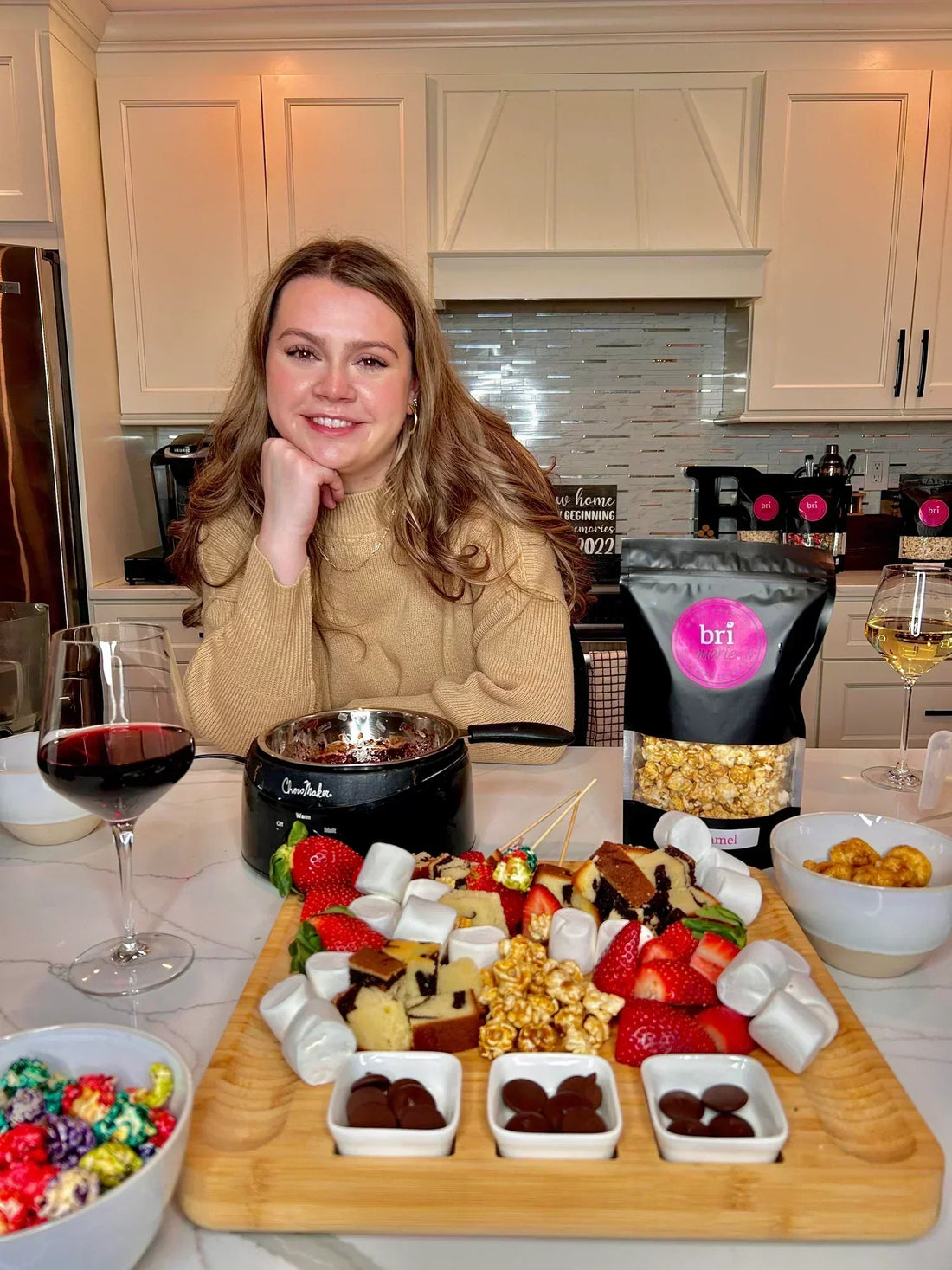 Bri Marie Popcorn founder seated in a kitchen beside a dessert board, wine, and assorted gourmet popcorn bags.