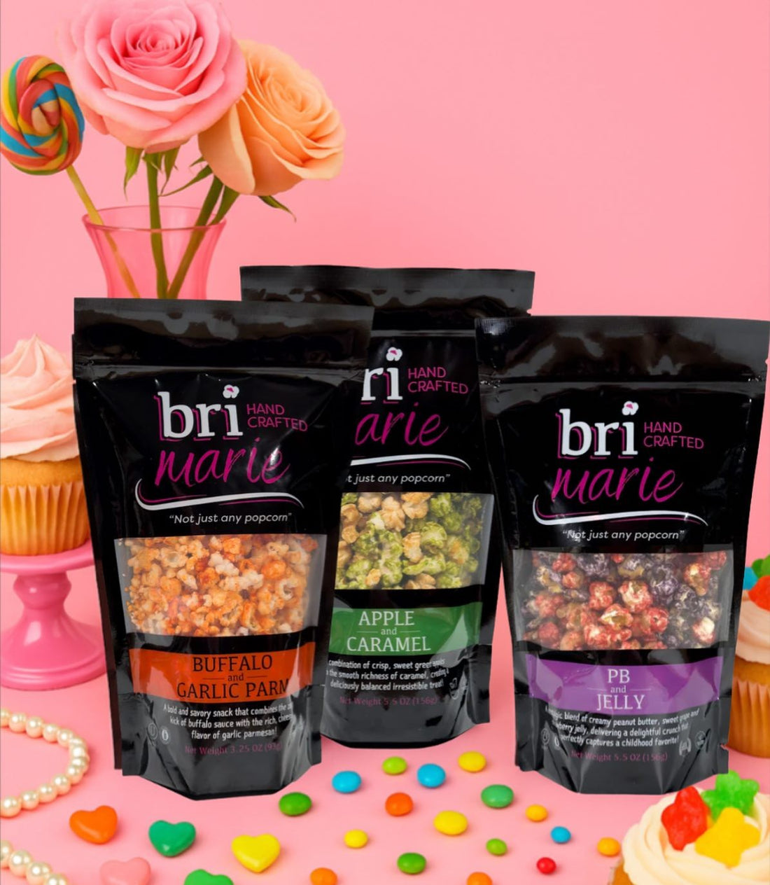 Three pouches of Bri Marie gourmet popcorn (Buffalo Garlic Parm, Apple Caramel, PB&J) on a pink background with candy.